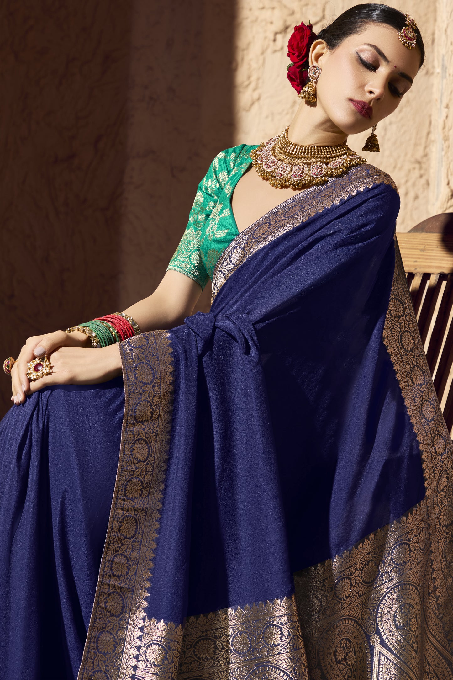 Closeup of a woman in a royal blue saree with gold embroidery and a green brocade blouse, adorned with a gold choker, jhumkas, bangles, and red roses in her hair. She sits elegantly, leaning slightly with soft lighting highlighting the fabric and jewelry.