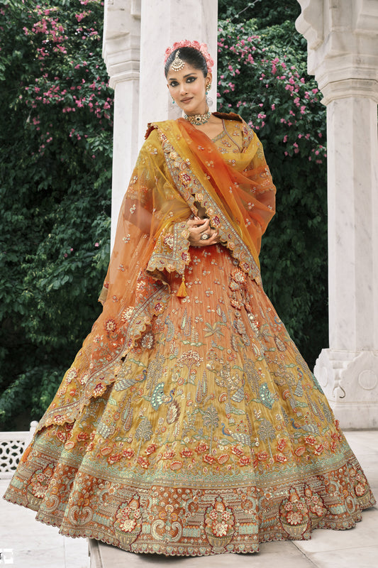 Model wearing a luxurious mustard-orange embroidered bridal lehenga adorned with intricate peacock and floral zari work, styled with traditional jewelry in a regal outdoor setting