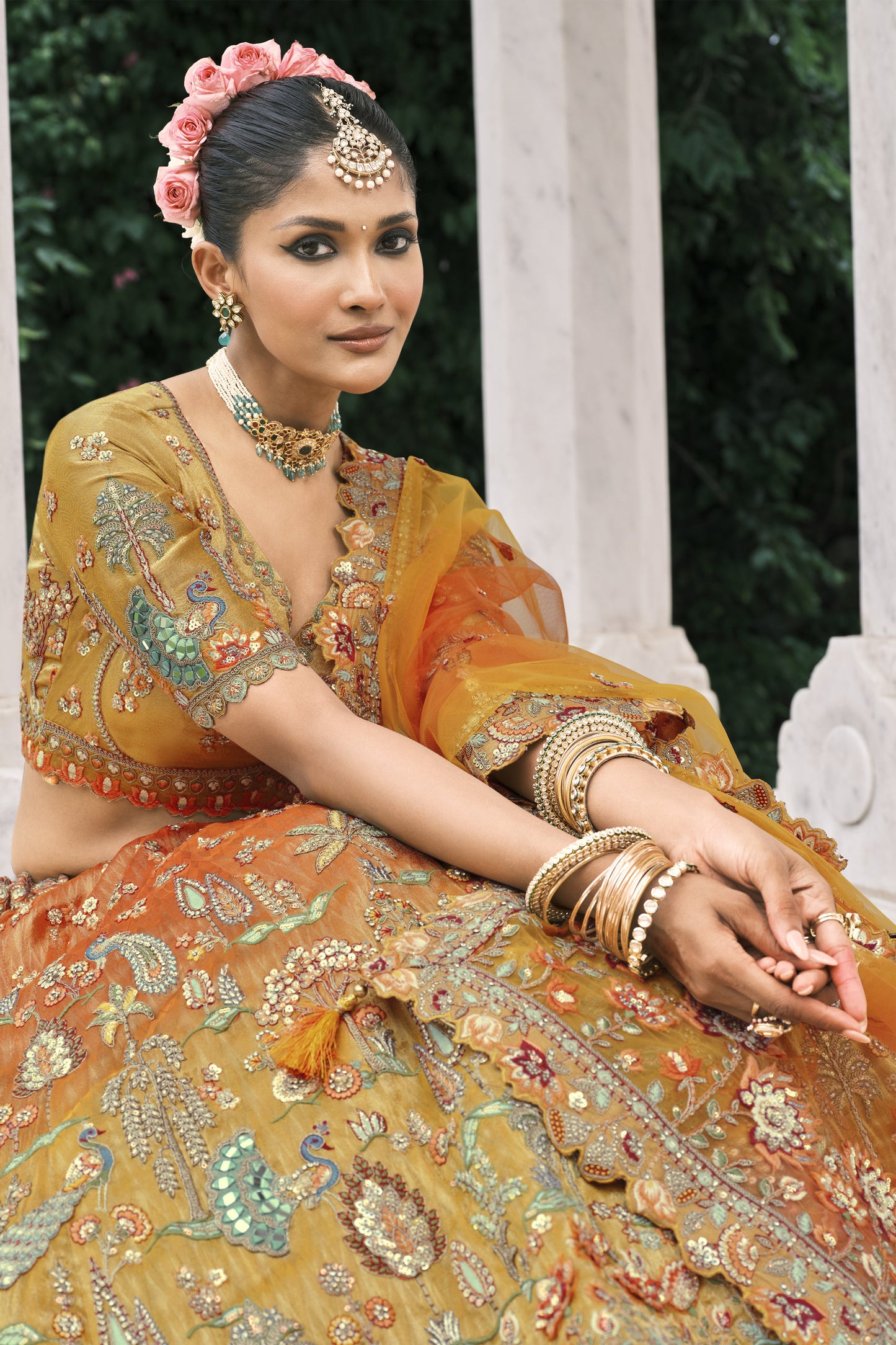Model wearing a luxury mustard-orange bridal lehenga with heavy zari embroidery, peacock motifs, sequins, and ornate dupatta in a royal architectural setting