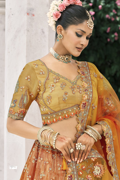 Model wearing a luxurious mustard-orange embroidered bridal lehenga adorned with intricate peacock and floral zari work, styled with traditional jewelry in a regal outdoor setting