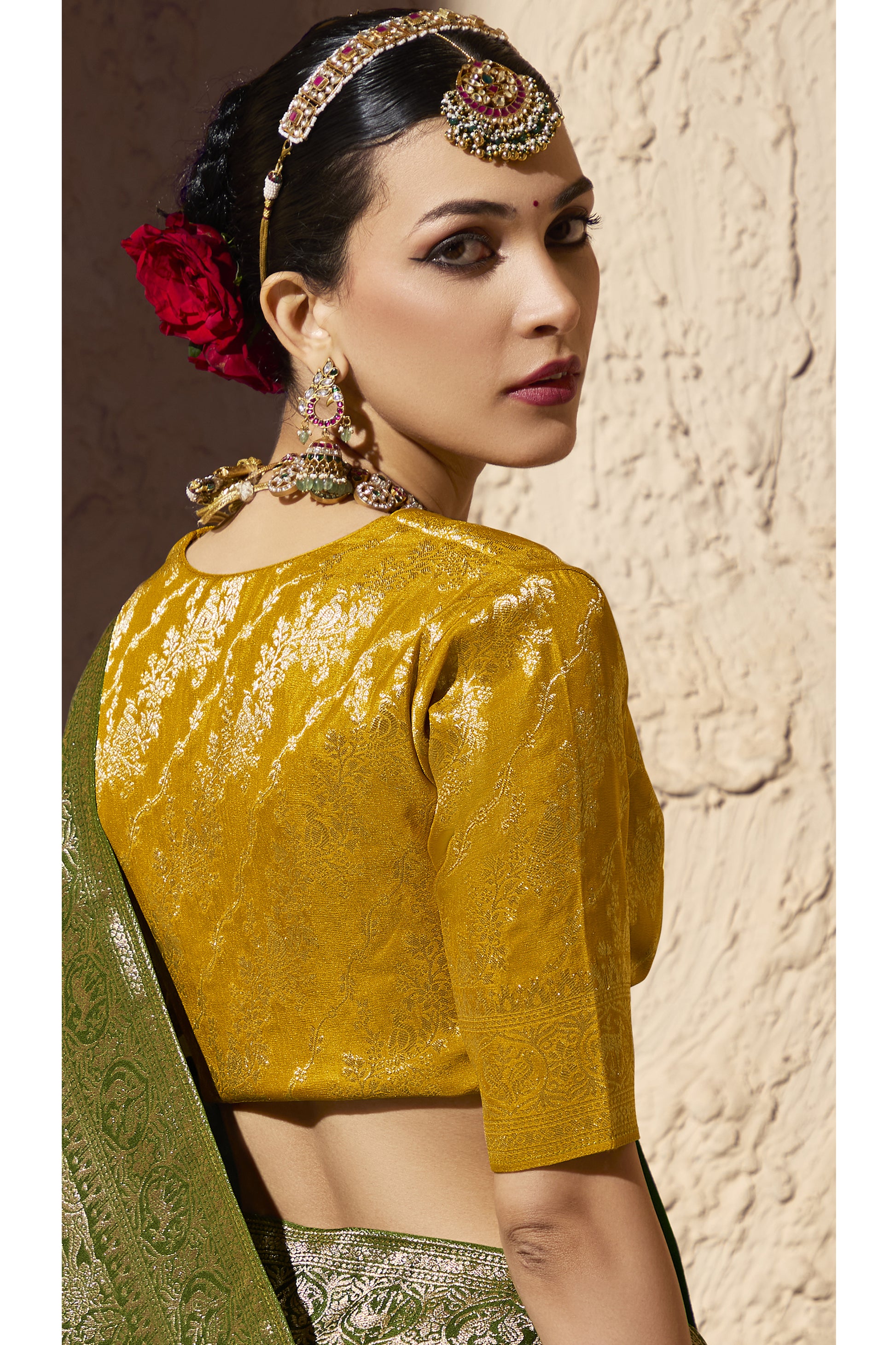 Green Banarasi-style silk saree with intricate gold embroidery paired with a mustard yellow brocade blouse, worn with layered pearl necklace, bangles, and heavy bridal jewelry.