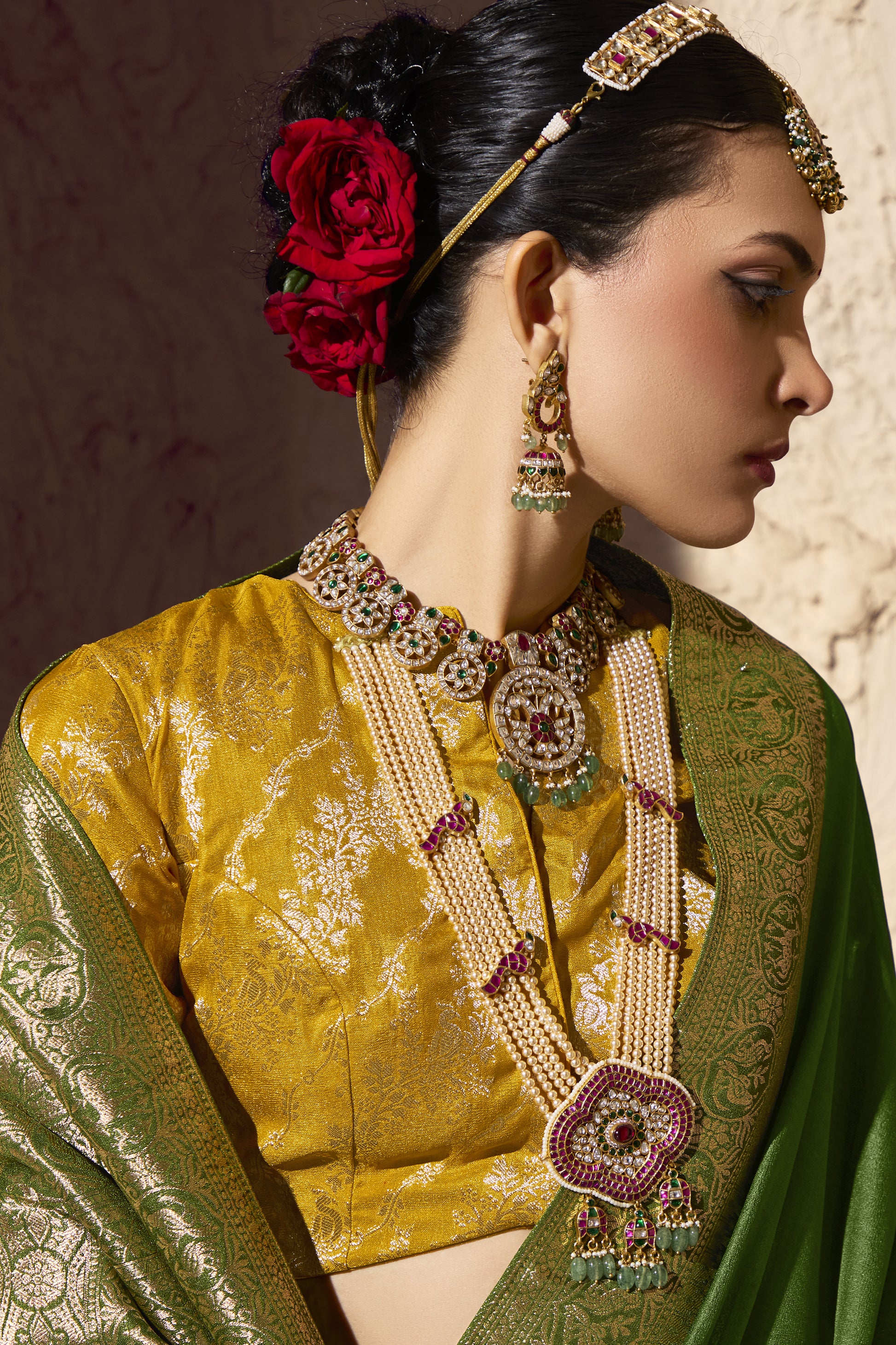 Close up of mustard yellow brocade blouse and green zari saree showing detailed gold embroidery, bridal jewelry with pearls, kundan stones, and red rose hair adornment.