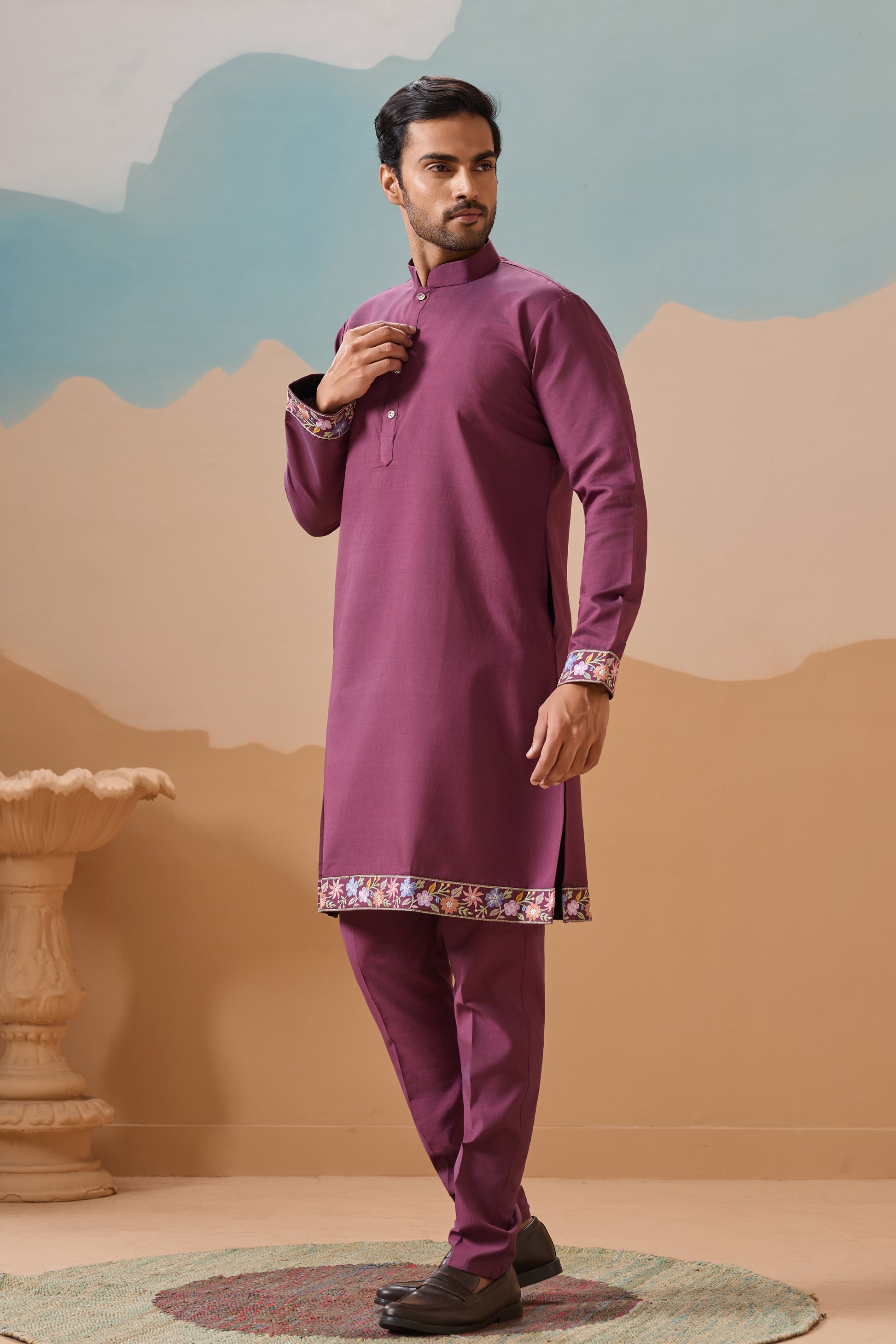 Man wearing a purple traditional outfit with a decorative hem against a stylized mountain background.
