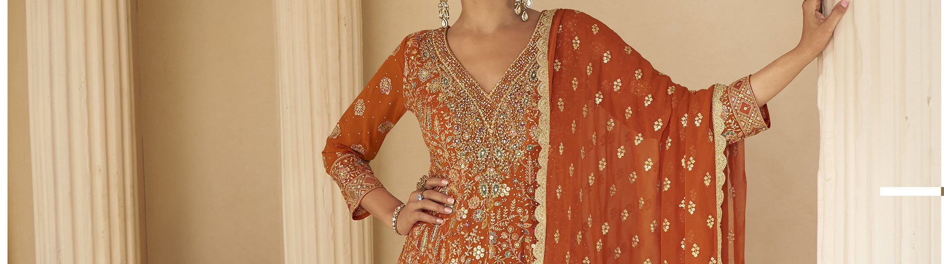Woman wearing a traditional orange and gold embroidered outfit against a beige curtain background


