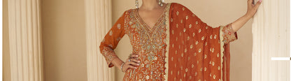 Woman wearing a traditional orange and gold embroidered outfit against a beige curtain background

