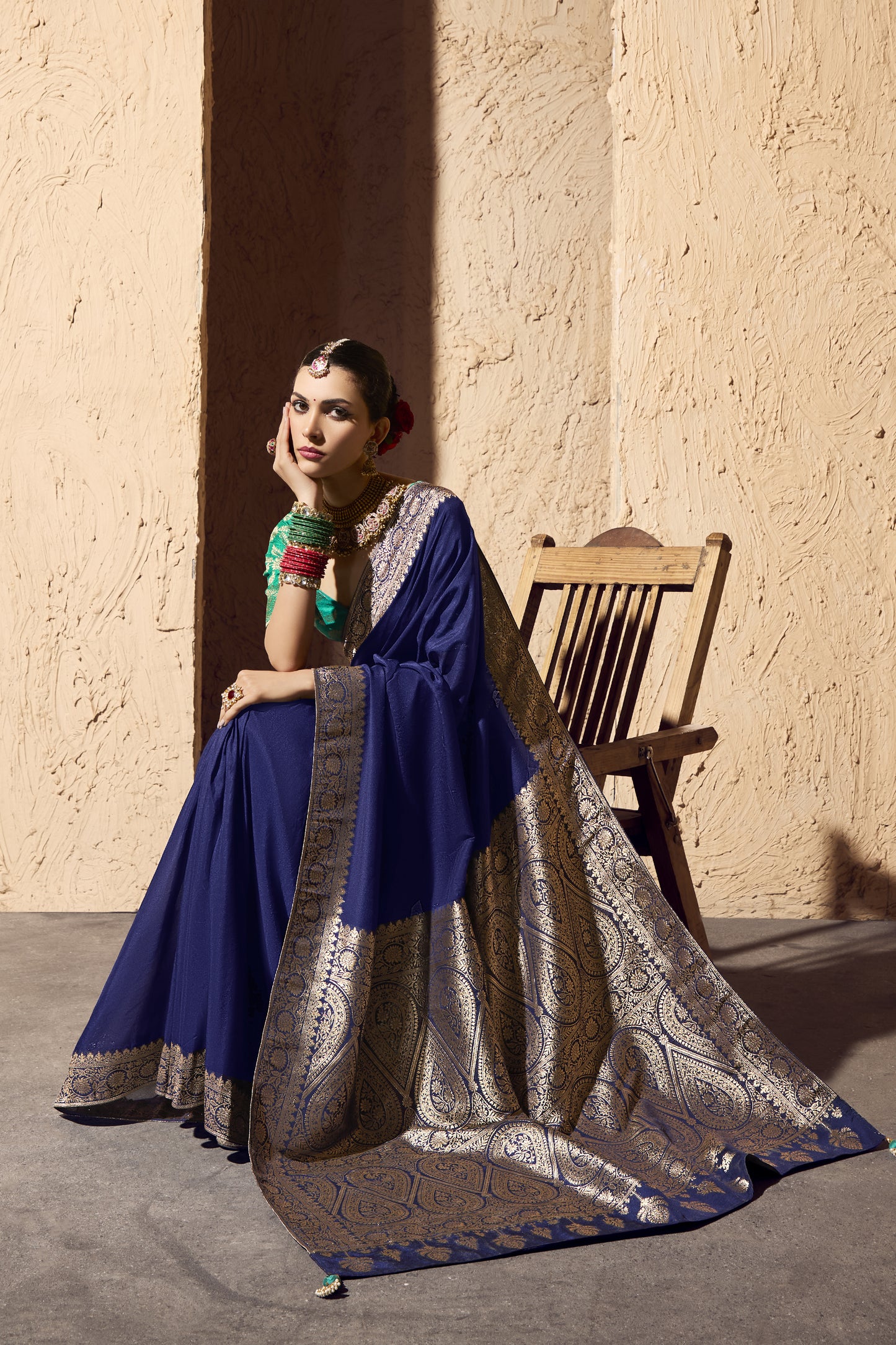 Woman seated gracefully in a royal blue saree with an ornate gold border, paired with a green brocade blouse. She wears heavy traditional gold jewelry, colorful bangles, and red flowers in her bun, posed against a textured beige wall beside a wooden chair.