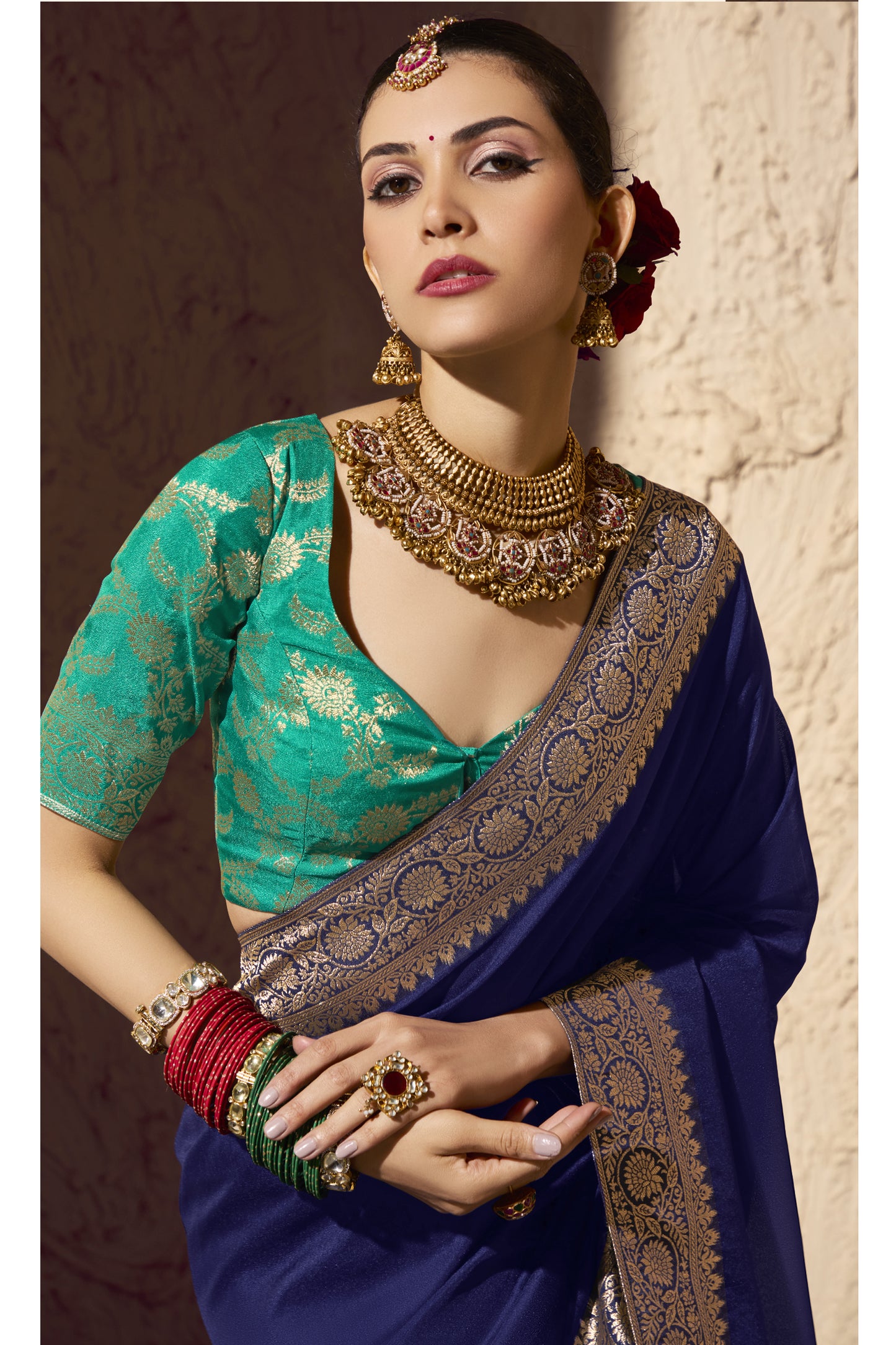 Close-up of a woman in a royal blue saree and green brocade blouse, adorned with a heavy gold choker, jhumka earrings, maang-tikka, and colorful bangles, showcasing a refined traditional Indian look against a softly lit textured background.