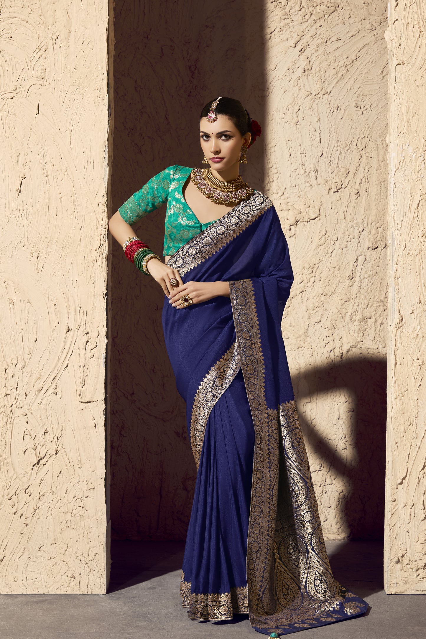 Woman wearing a royal blue saree with an intricate gold border and a green brocade blouse, accessorized with traditional gold jewelry, bangles, and a sleek bun with red flowers, posing elegantly against a textured beige backdrop